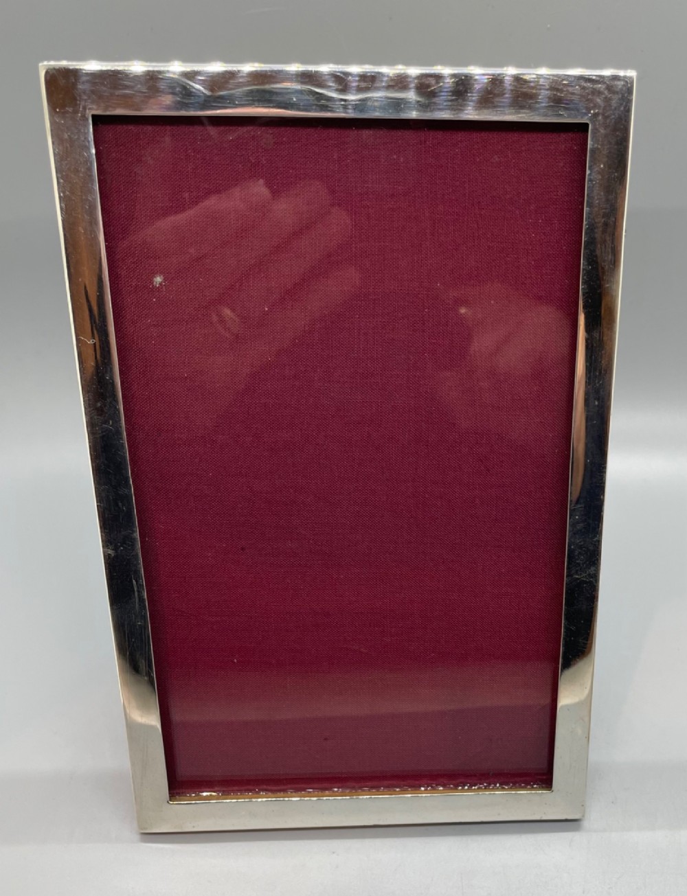 An upright rectangular sterling silver photo frame with a polished border, easel back stand, and burgundy fabric interior. View II