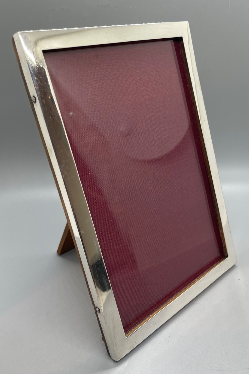 An upright rectangular sterling silver photo frame with a polished border, easel back stand, and burgundy fabric interior. View IX