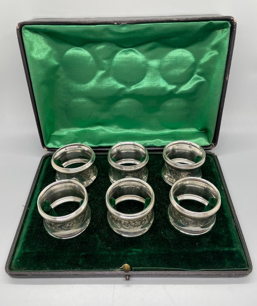 A set of six circular Edwardian sterling silver napkin rings with engraved patterns, displayed inside an open presentation box with green velvet lining. View II