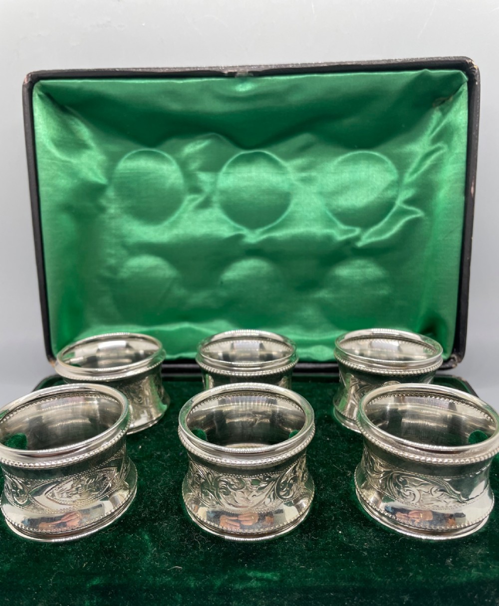 A set of six circular Edwardian sterling silver napkin rings with engraved patterns, displayed inside an open presentation box with green velvet lining. View III