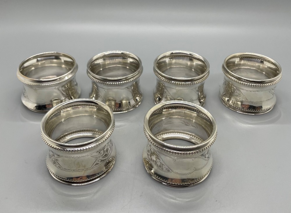A set of six circular Edwardian sterling silver napkin rings with engraved patterns, displayed inside an open presentation box with green velvet lining. View IV