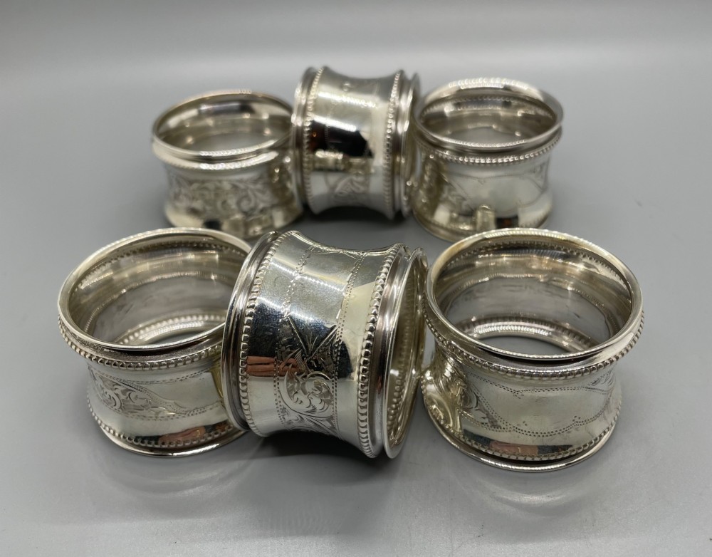 A set of six circular Edwardian sterling silver napkin rings with engraved patterns, displayed inside an open presentation box with green velvet lining. View V