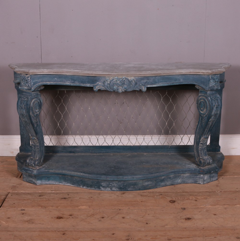 A French-style serpentine console table featuring a distressed blue painted finish, ornate carved scroll supports, a lower shelf, and a wire mesh back panel.