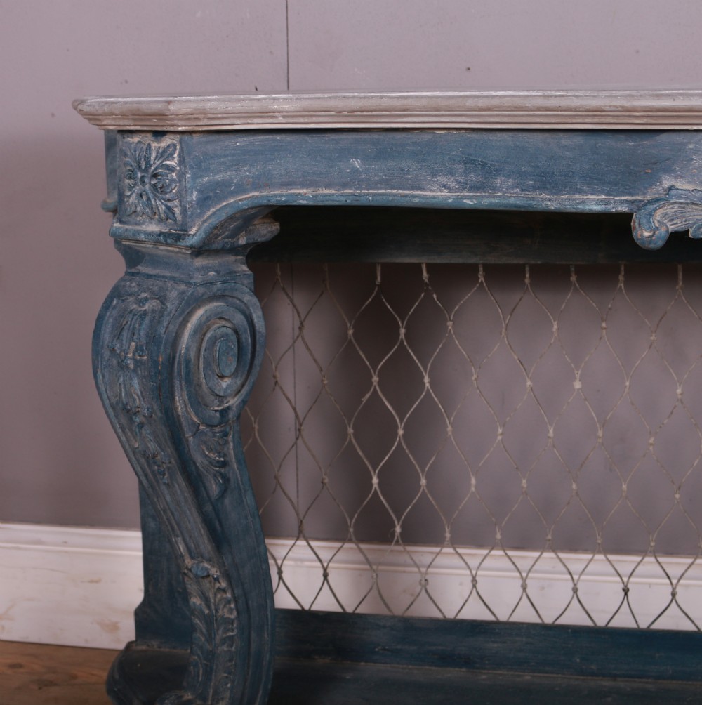 A French-style serpentine console table featuring a distressed blue painted finish, ornate carved scroll supports, a lower shelf, and a wire mesh back panel. View III
