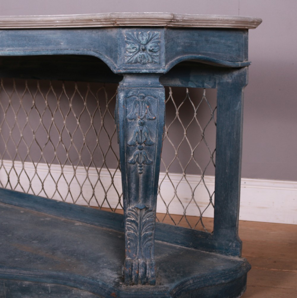 A French-style serpentine console table featuring a distressed blue painted finish, ornate carved scroll supports, a lower shelf, and a wire mesh back panel. View IV