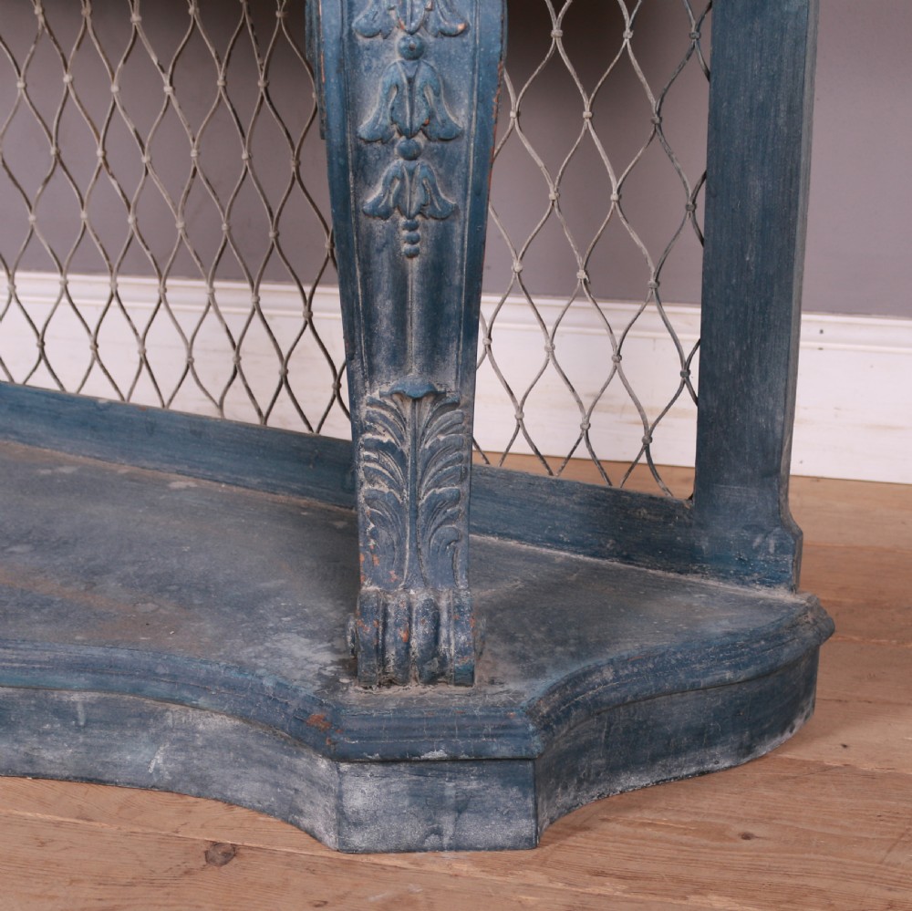 A French-style serpentine console table featuring a distressed blue painted finish, ornate carved scroll supports, a lower shelf, and a wire mesh back panel. View V