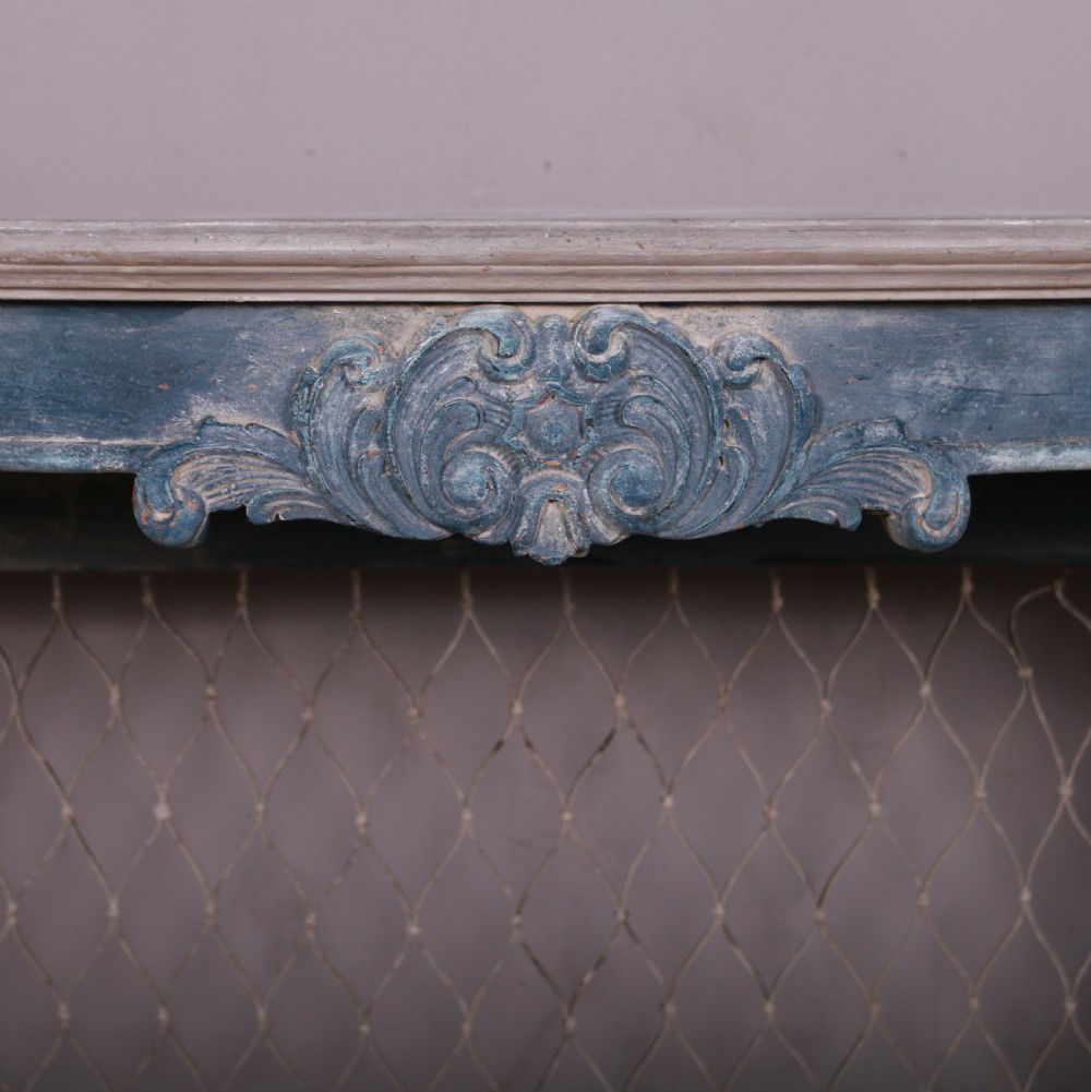 A French-style serpentine console table featuring a distressed blue painted finish, ornate carved scroll supports, a lower shelf, and a wire mesh back panel. View VI