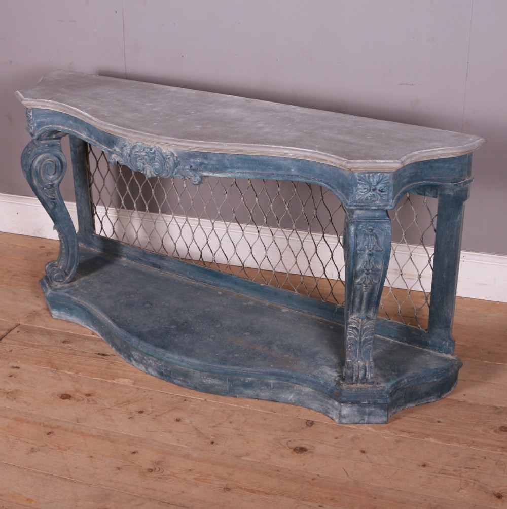A French-style serpentine console table featuring a distressed blue painted finish, ornate carved scroll supports, a lower shelf, and a wire mesh back panel. View VII