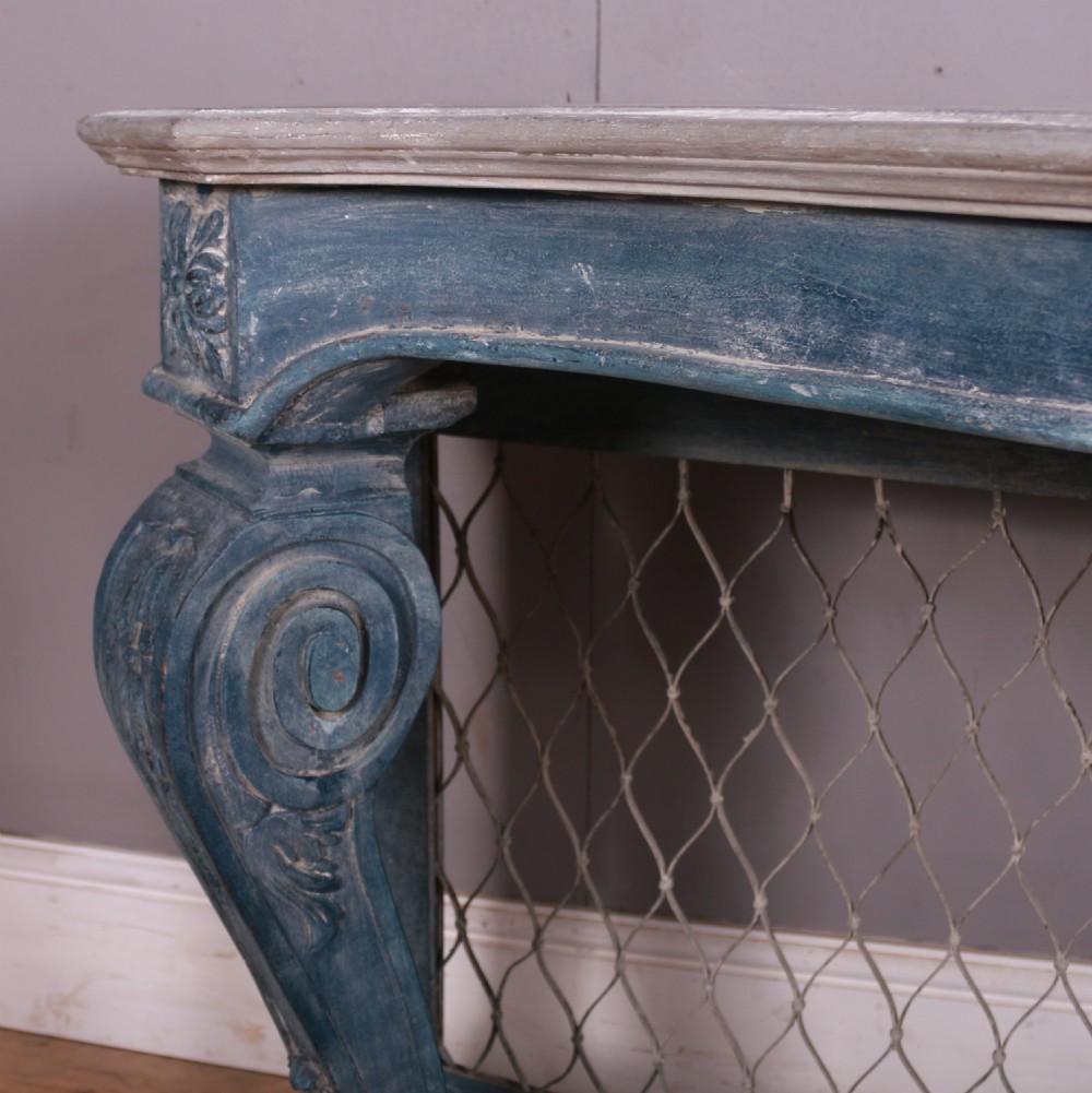 A French-style serpentine console table featuring a distressed blue painted finish, ornate carved scroll supports, a lower shelf, and a wire mesh back panel. View VIII