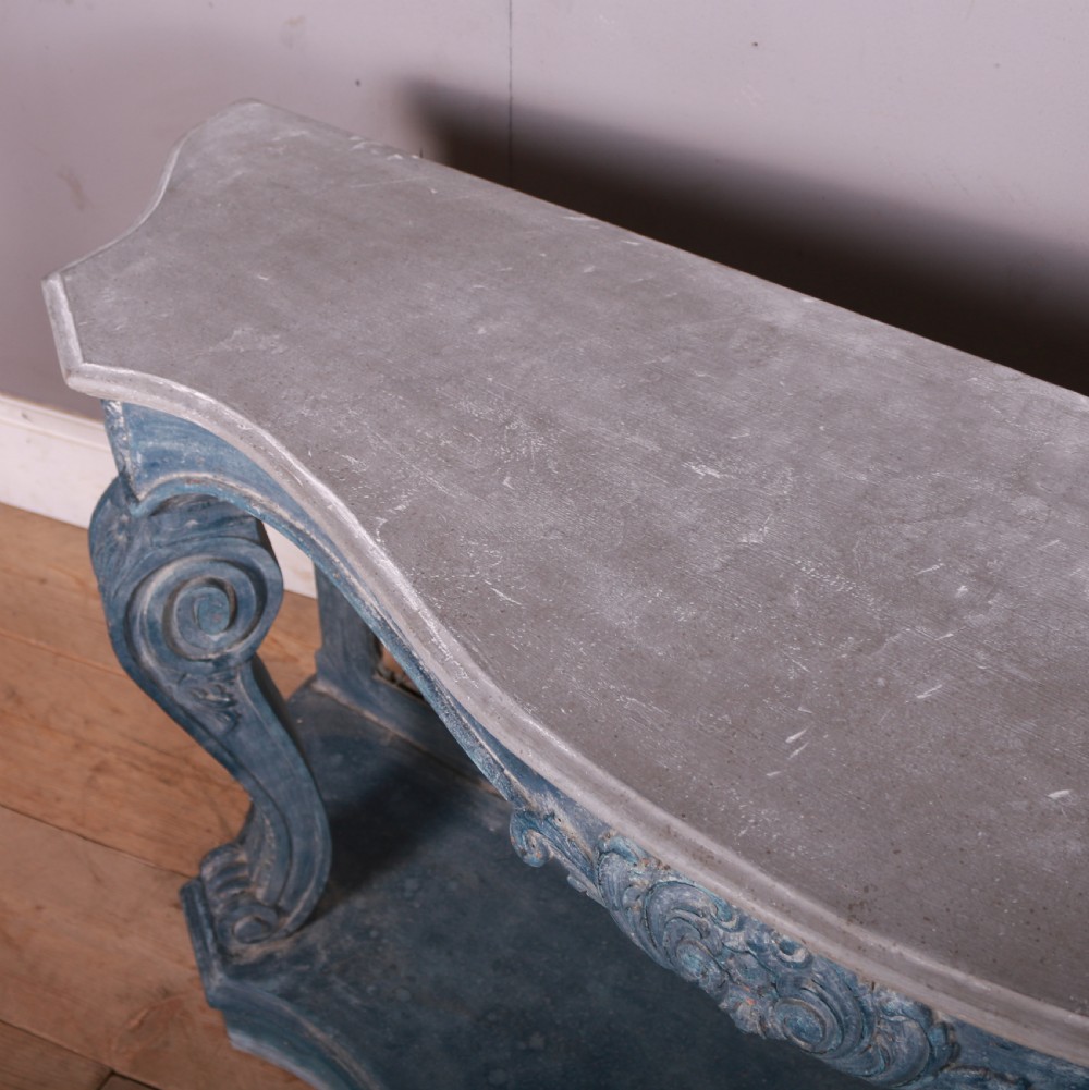 A French-style serpentine console table featuring a distressed blue painted finish, ornate carved scroll supports, a lower shelf, and a wire mesh back panel. View IX
