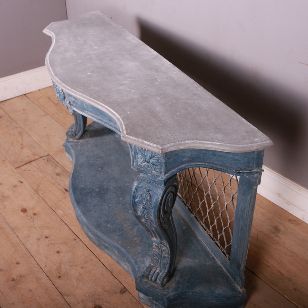 A French-style serpentine console table featuring a distressed blue painted finish, ornate carved scroll supports, a lower shelf, and a wire mesh back panel. View X