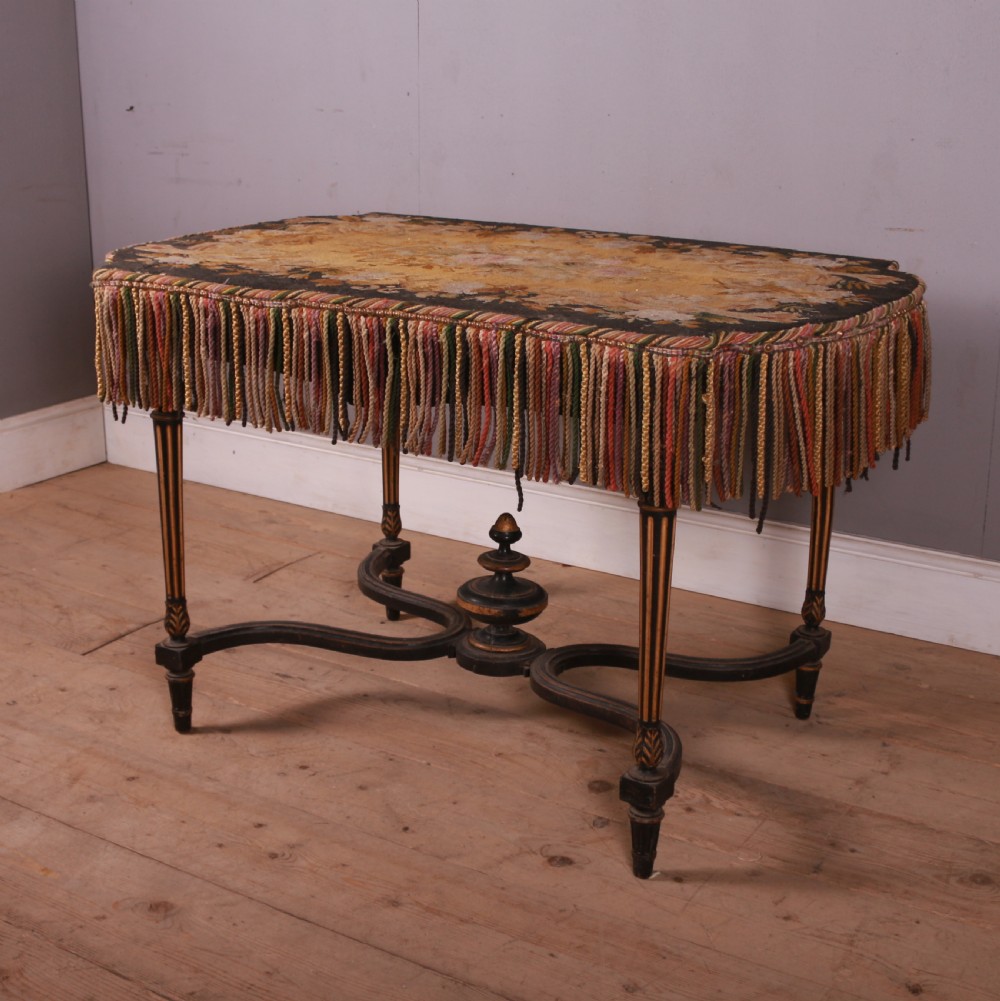 A 19th-century French ebonised centre table featuring fluted legs with gilt accents, a curved stretcher with a turned finial, and a floral tapestry top surrounded by long fringe.