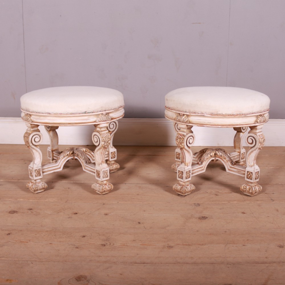 A pair of circular Swedish stools featuring cream and gold painted frames, scroll legs, arched stretchers, and off-white upholstered seats.