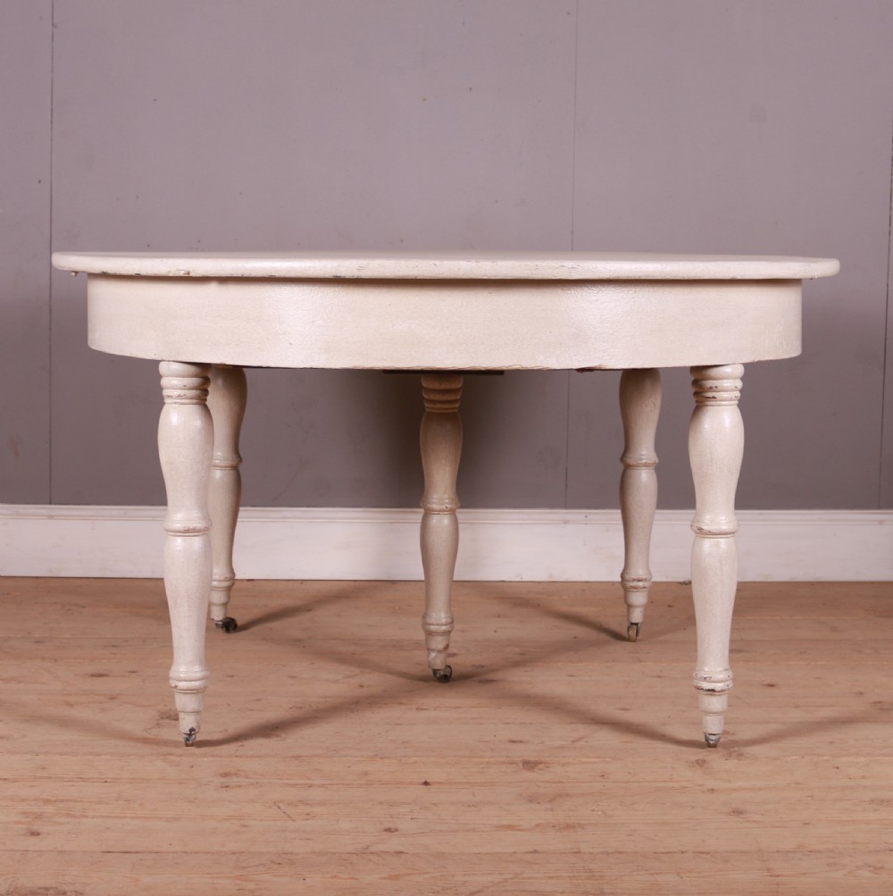 A circular off-white painted wooden dining table featuring five turned legs fitted with brass castors.