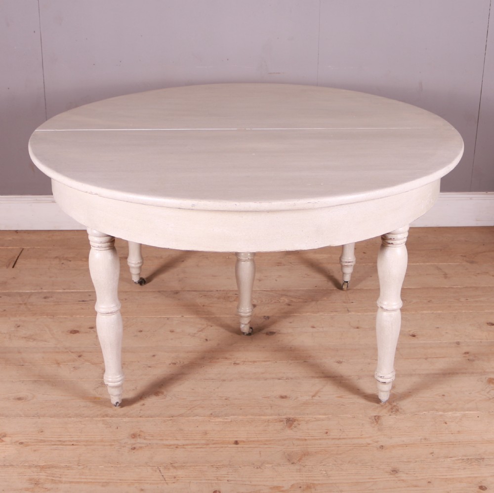 A circular off-white painted wooden dining table featuring five turned legs fitted with brass castors. View II