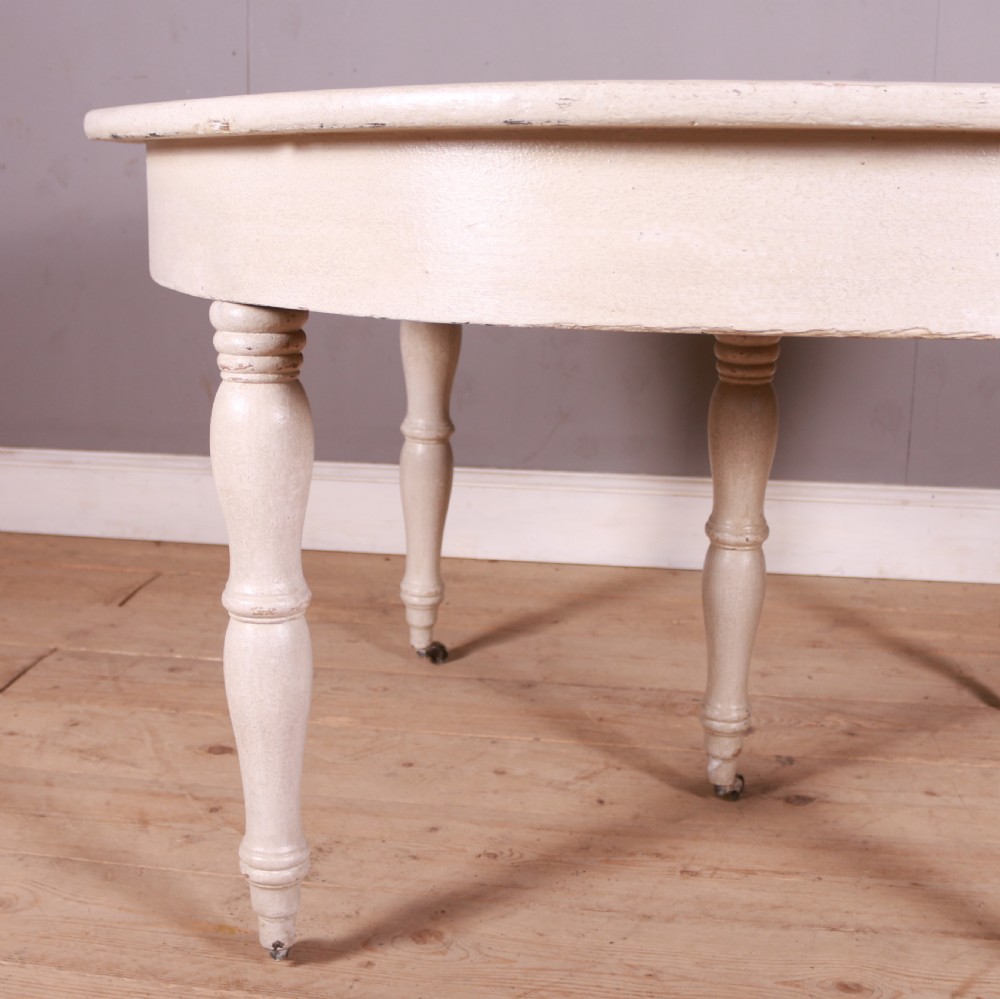 A circular off-white painted wooden dining table featuring five turned legs fitted with brass castors. View III
