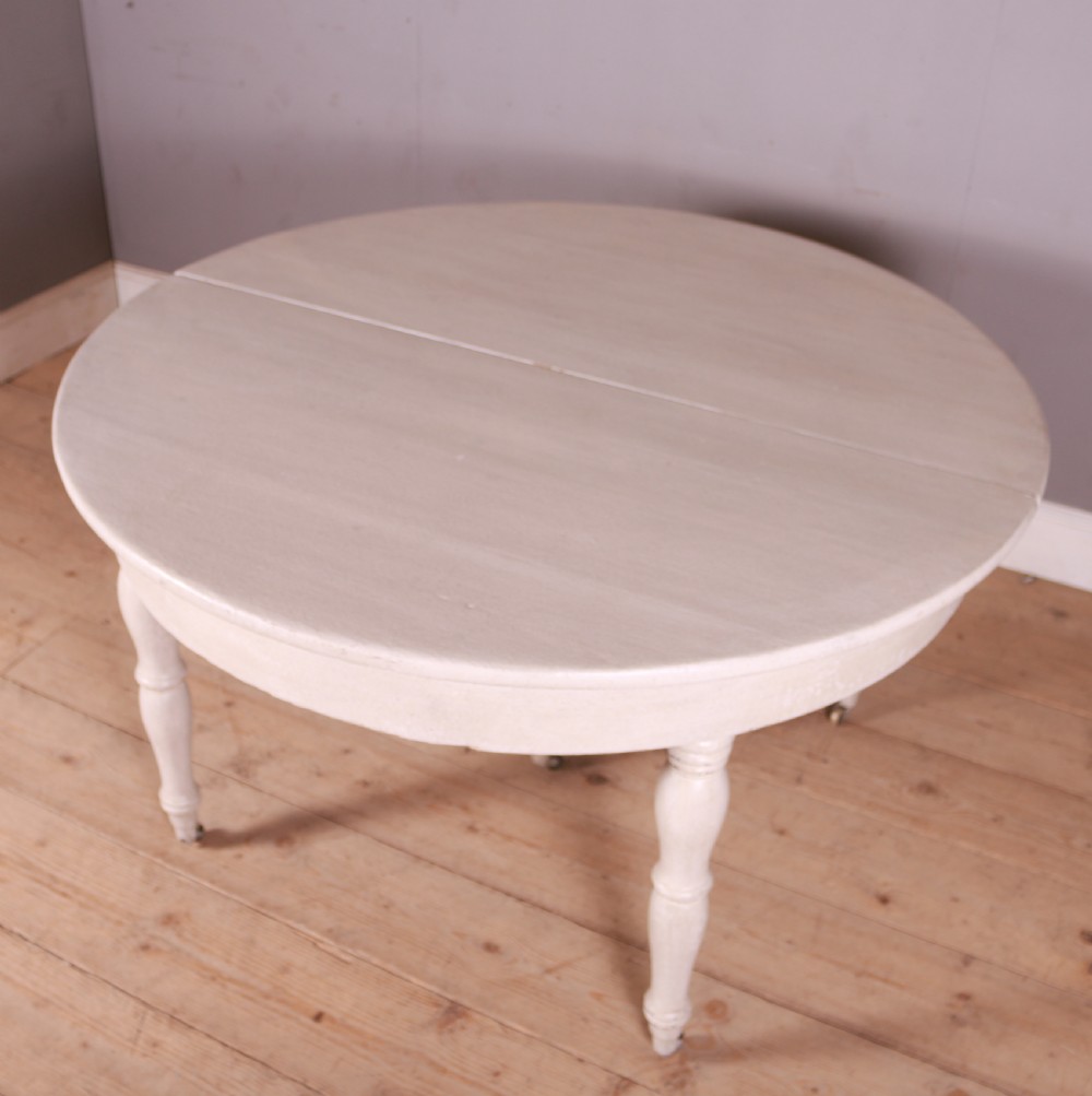 A circular off-white painted wooden dining table featuring five turned legs fitted with brass castors. View IV