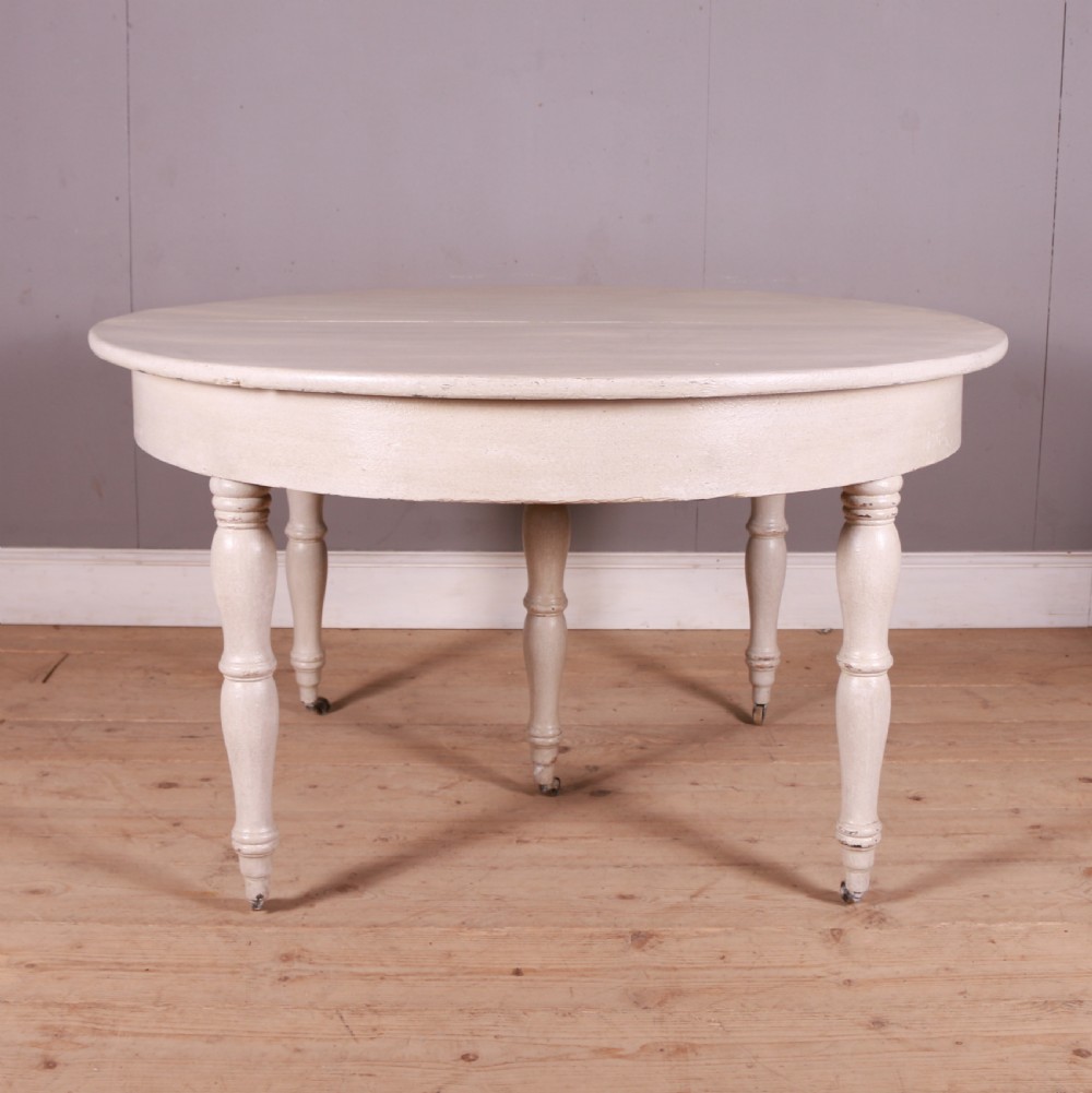 A circular off-white painted wooden dining table featuring five turned legs fitted with brass castors. View V