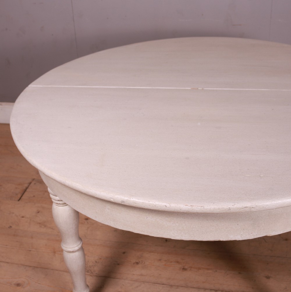 A circular off-white painted wooden dining table featuring five turned legs fitted with brass castors. View VI