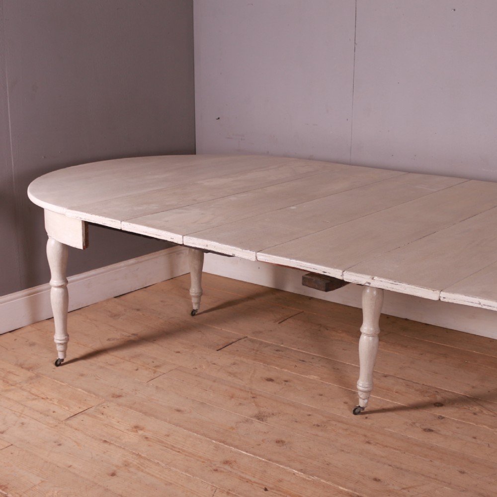 A circular off-white painted wooden dining table featuring five turned legs fitted with brass castors. View VIII