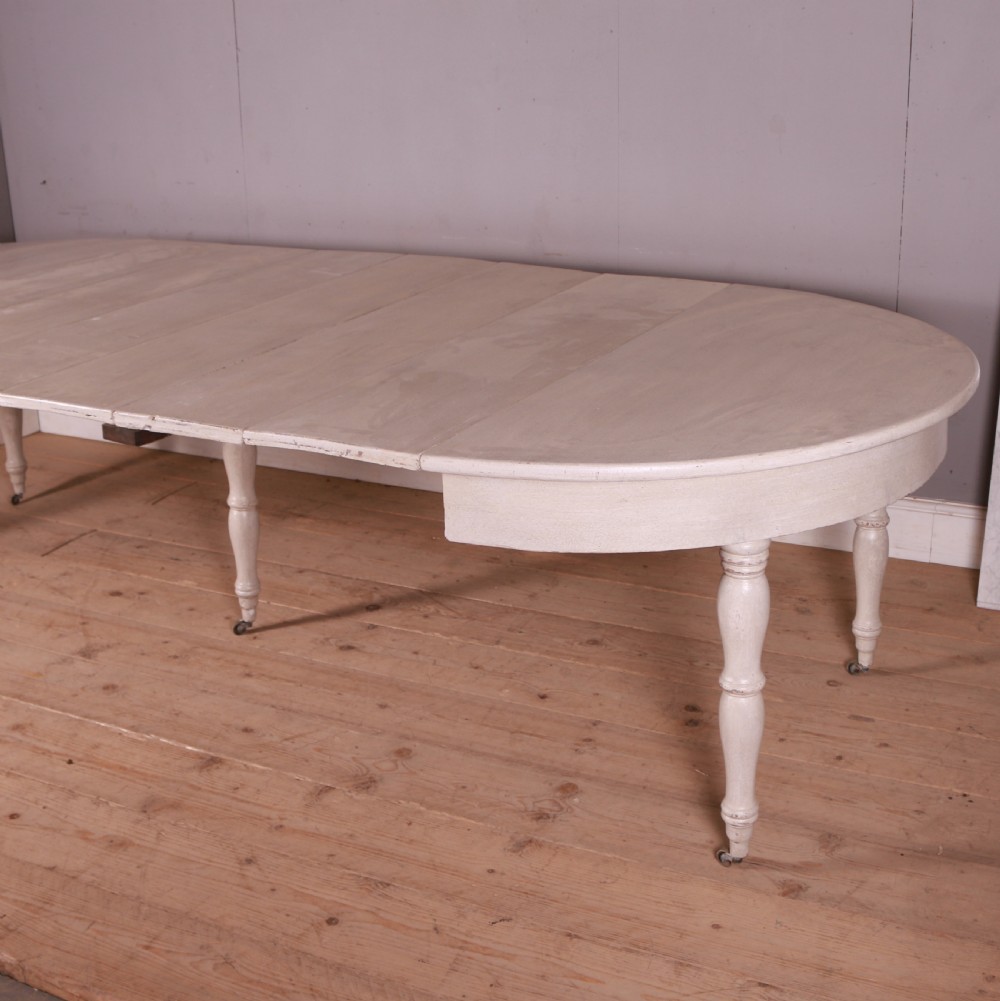 A circular off-white painted wooden dining table featuring five turned legs fitted with brass castors. View IX