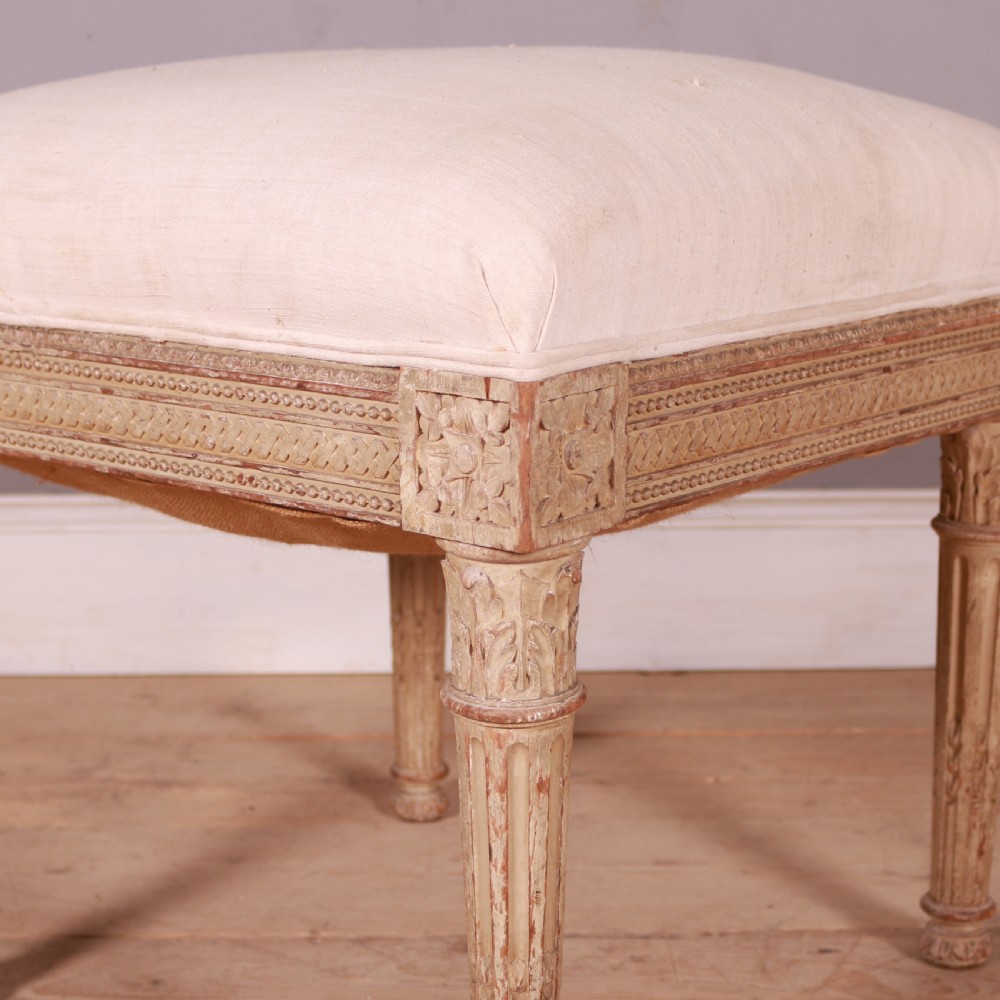 A square Swedish Gustavian upholstered stool featuring distressed cream painted wood legs with fluted carving and a light linen seat. View II