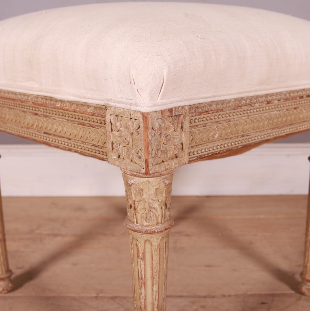 A square Swedish Gustavian upholstered stool featuring distressed cream painted wood legs with fluted carving and a light linen seat. View V