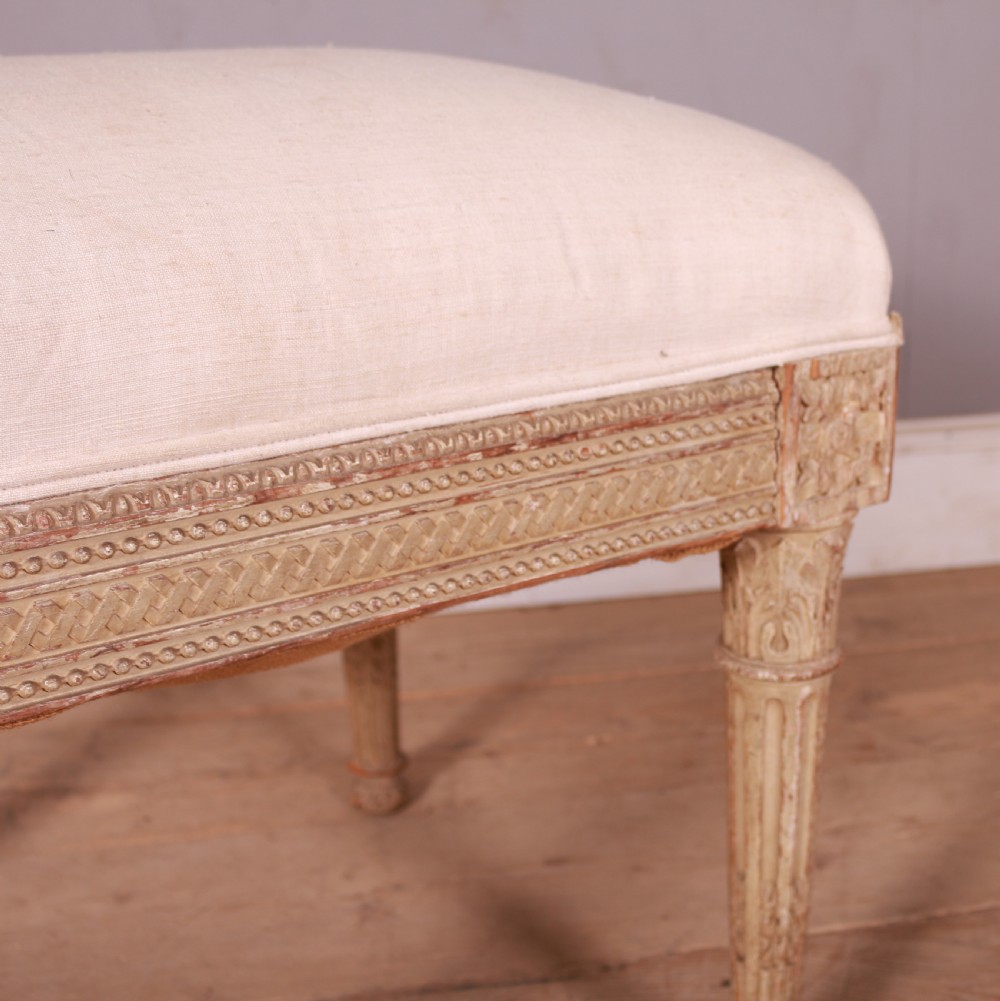 A square Swedish Gustavian upholstered stool featuring distressed cream painted wood legs with fluted carving and a light linen seat. View VI