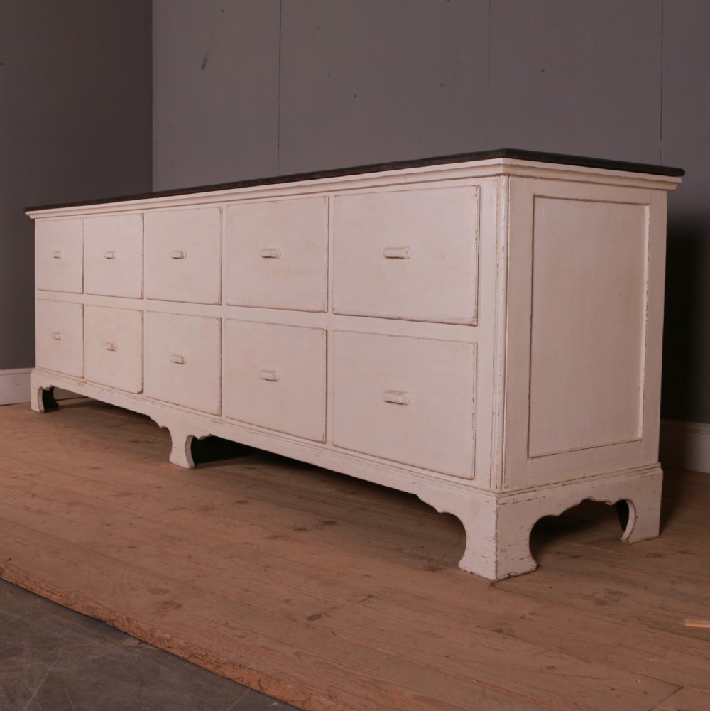A long off-white painted French bank of drapers drawers featuring ten large drawers with wooden handles and a dark stained top surface.