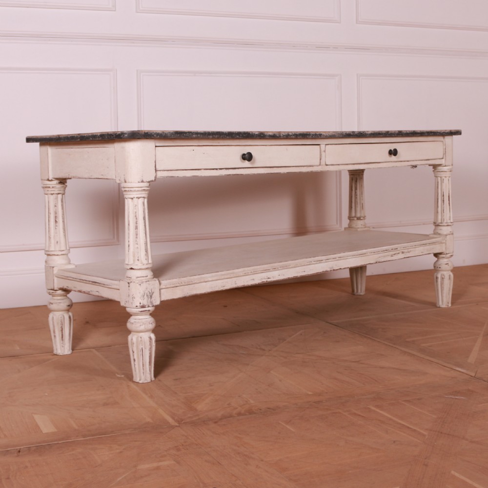 A cream painted pine side table featuring two drawers, turned fluted legs, a lower open shelf, and a dark contrasting top.