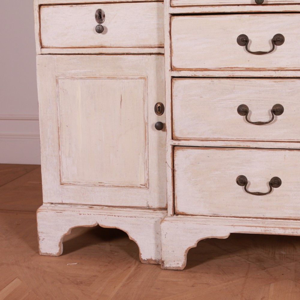 An eighteenth-century off-white painted housekeepers cupboard featuring fielded panel cupboard doors above multiple drawers with brass handles, supported by bracket feet. View VII