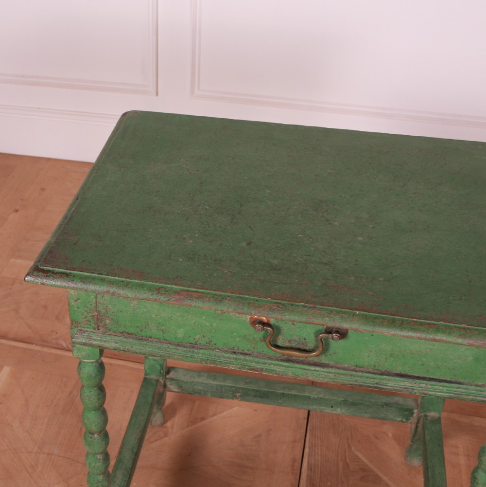 An 18th-century green painted wooden lamp table featuring four bobbin-turned legs, a rectangular top, lower stretchers, and a single drawer with a brass drop handle. View VI