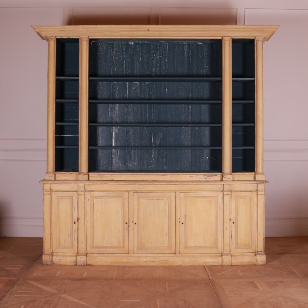 A large antique French painted bookcase featuring a pale yellow exterior, open shelving with a dark blue painted interior, and panelled cupboard doors at the base.