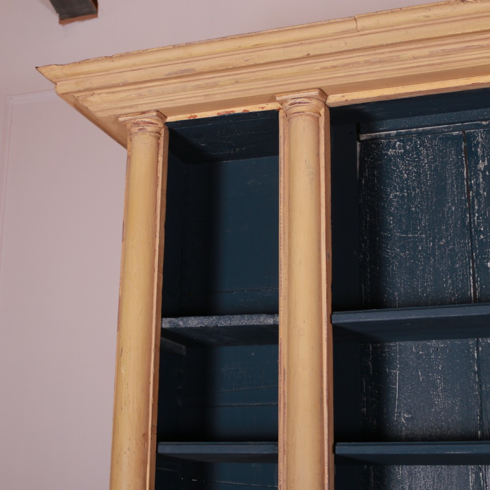 A large antique French painted bookcase featuring a pale yellow exterior, open shelving with a dark blue painted interior, and panelled cupboard doors at the base. View V