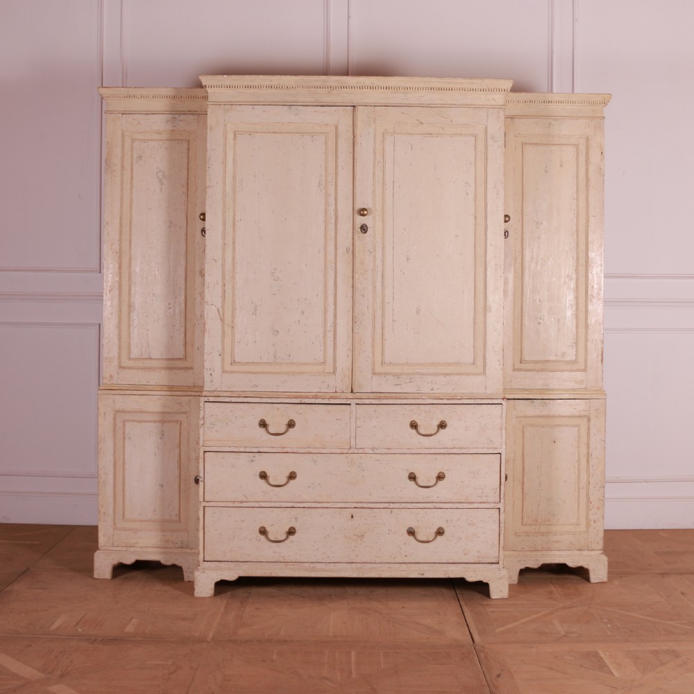 An antique cream-painted breakfront housekeepers cupboard featuring central panelled doors above drawers, flanked by tall side cupboards.