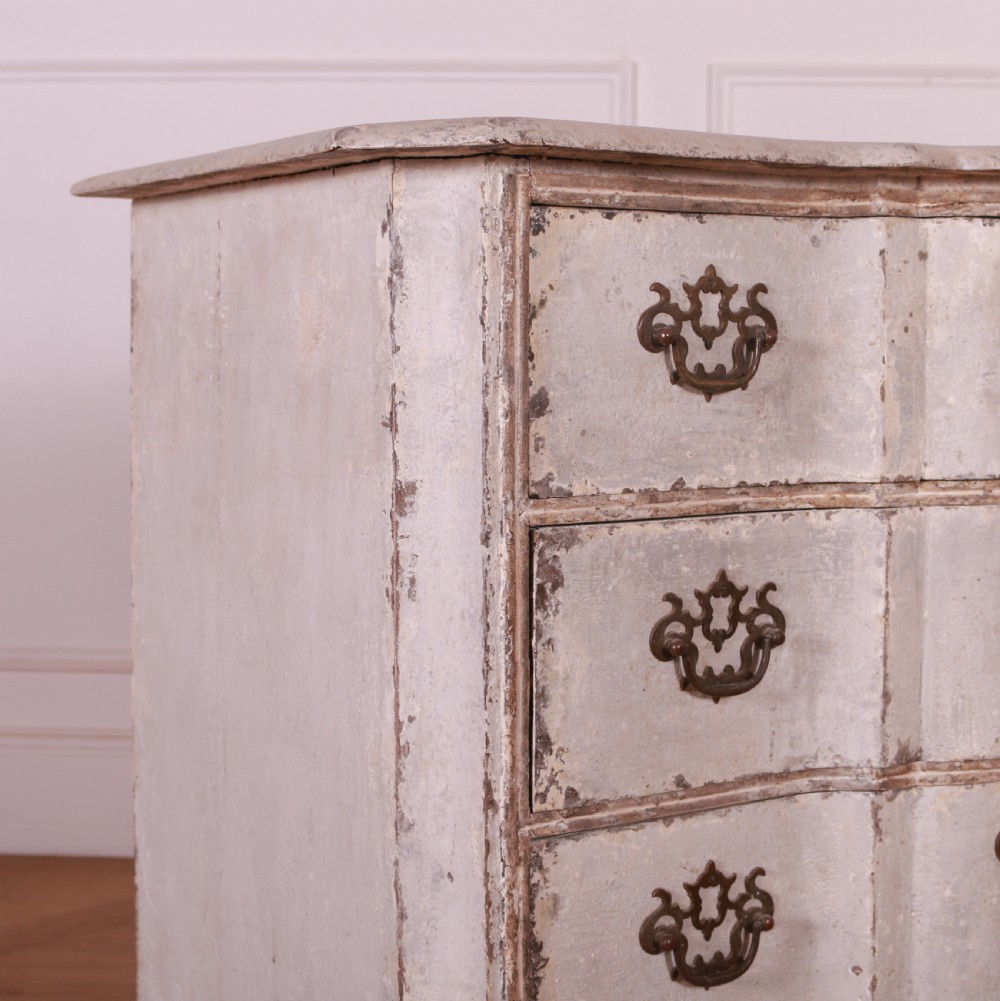 An 18th-century Dutch serpentine-front commode featuring four drawers with a distressed pale paint finish and ornate brass handles. View IV