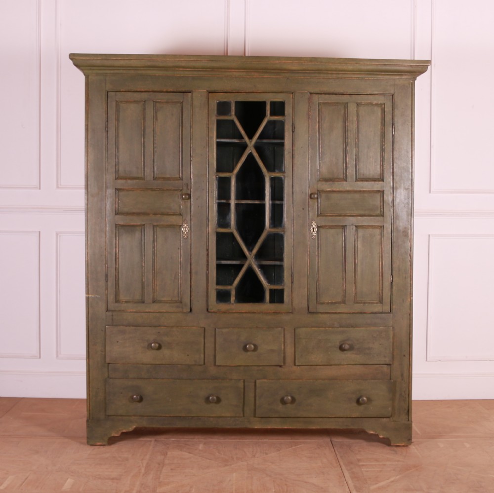 A large green painted pine cupboard featuring a central glazed door with geometric glazing bars flanked by two panelled doors above five drawers.