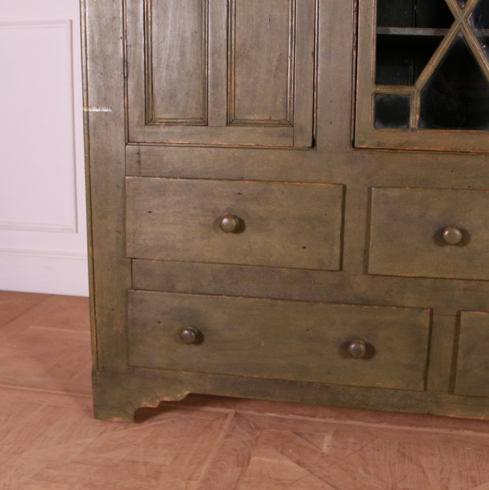 A large green painted pine cupboard featuring a central glazed door with geometric glazing bars flanked by two panelled doors above five drawers. View III