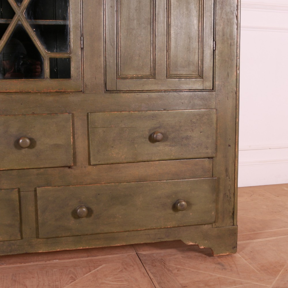 A large green painted pine cupboard featuring a central glazed door with geometric glazing bars flanked by two panelled doors above five drawers. View VI