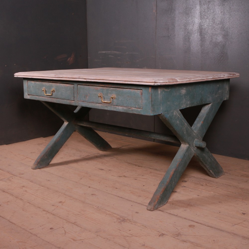 An antique English trestle table featuring a muted teal painted X-frame base, two apron drawers with brass handles, and a scrubbed natural pine top.