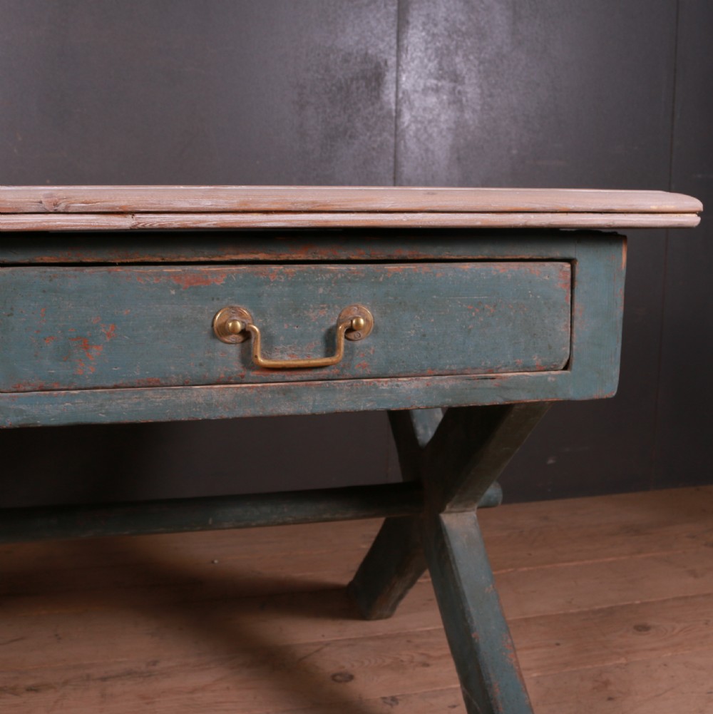 An antique English trestle table featuring a muted teal painted X-frame base, two apron drawers with brass handles, and a scrubbed natural pine top. View V