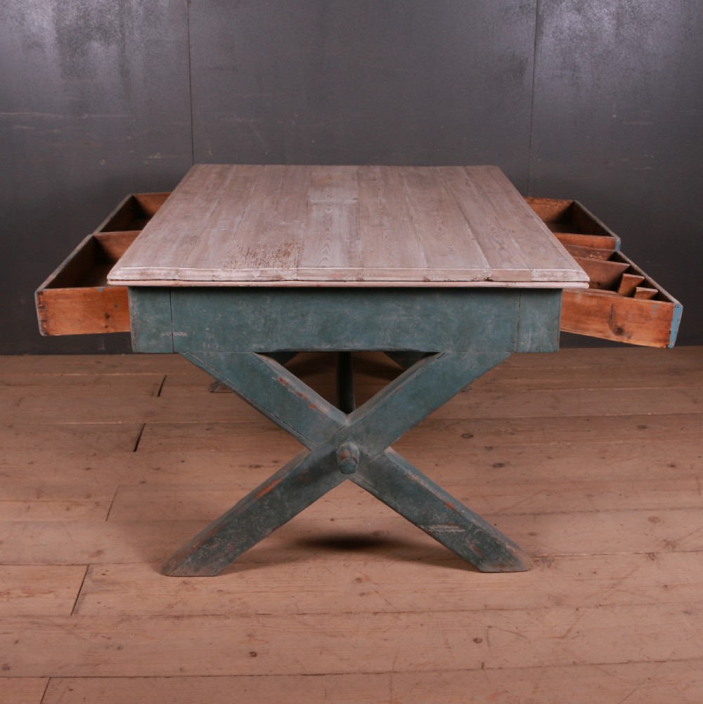 An antique English trestle table featuring a muted teal painted X-frame base, two apron drawers with brass handles, and a scrubbed natural pine top. View X