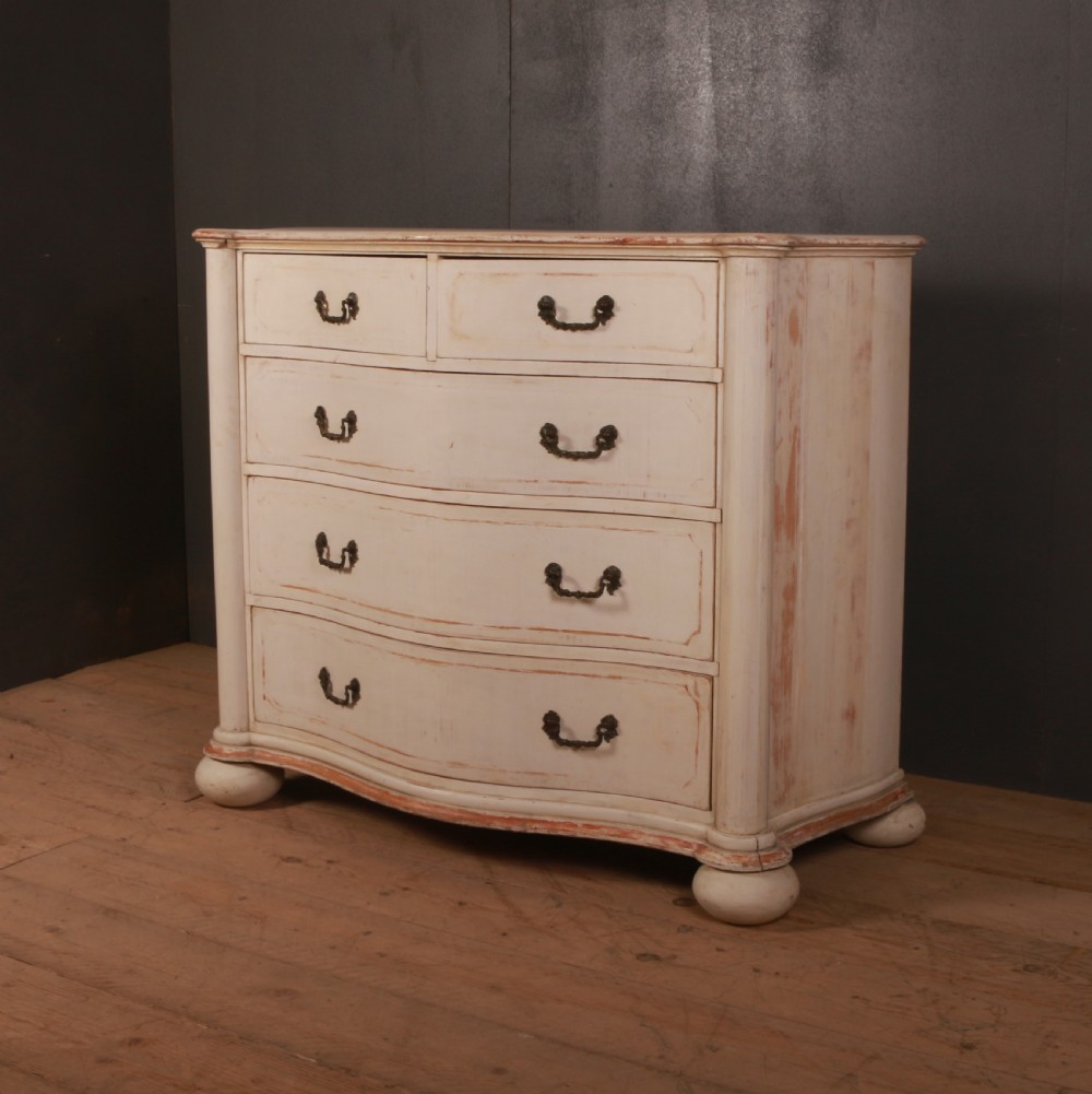 An antique cream painted Italian commode with a serpentine curved front, metal drop handles, and turned bun feet on a wood floor.