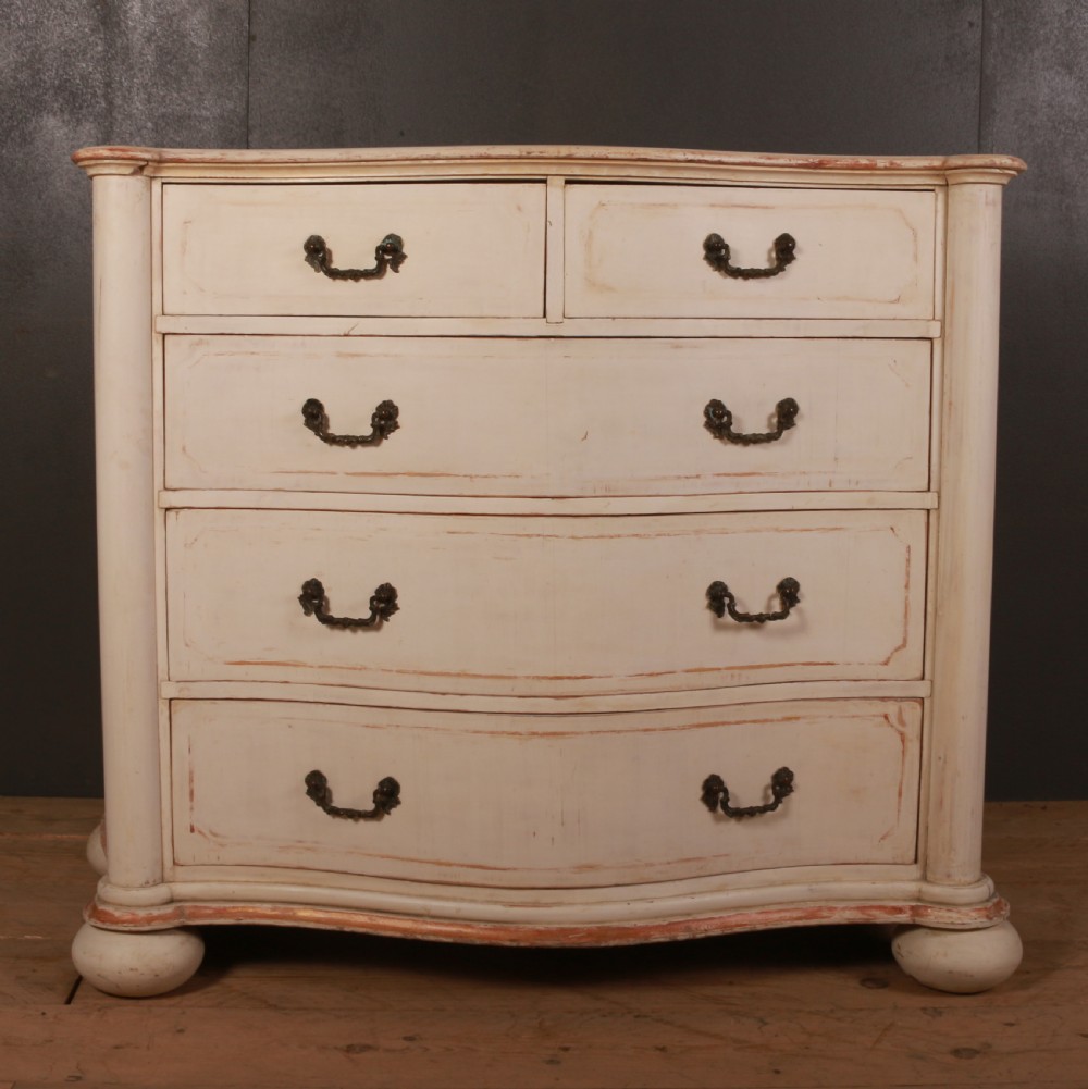 An antique cream painted Italian commode with a serpentine curved front, metal drop handles, and turned bun feet on a wood floor. View II