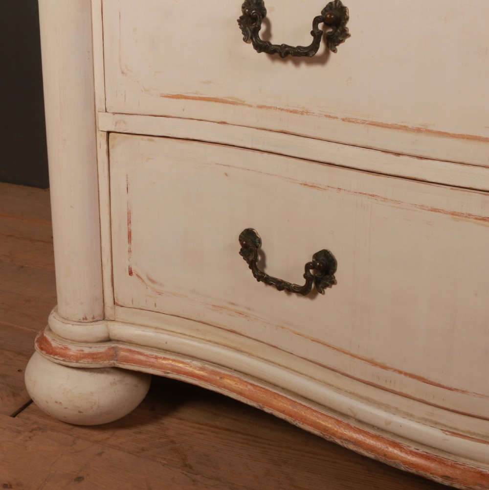 An antique cream painted Italian commode with a serpentine curved front, metal drop handles, and turned bun feet on a wood floor. View V