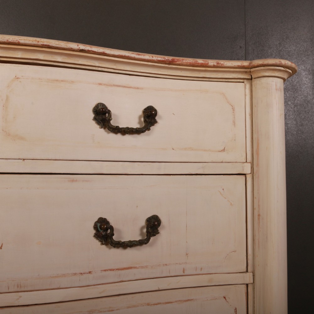 An antique cream painted Italian commode with a serpentine curved front, metal drop handles, and turned bun feet on a wood floor. View VI