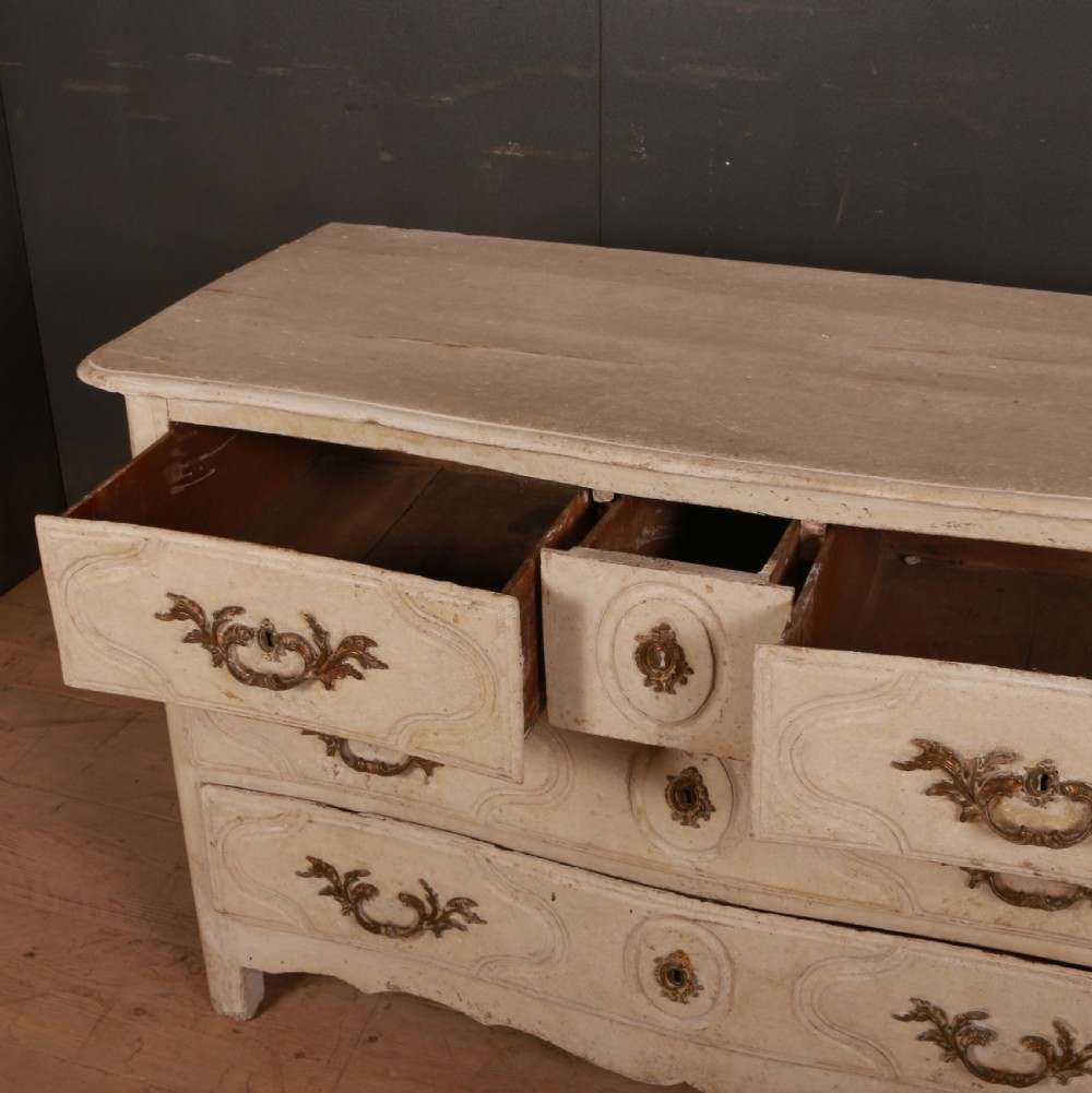 An off-white painted wooden French commode featuring three drawers with ornamental brass handles and a scalloped bottom apron. View VII