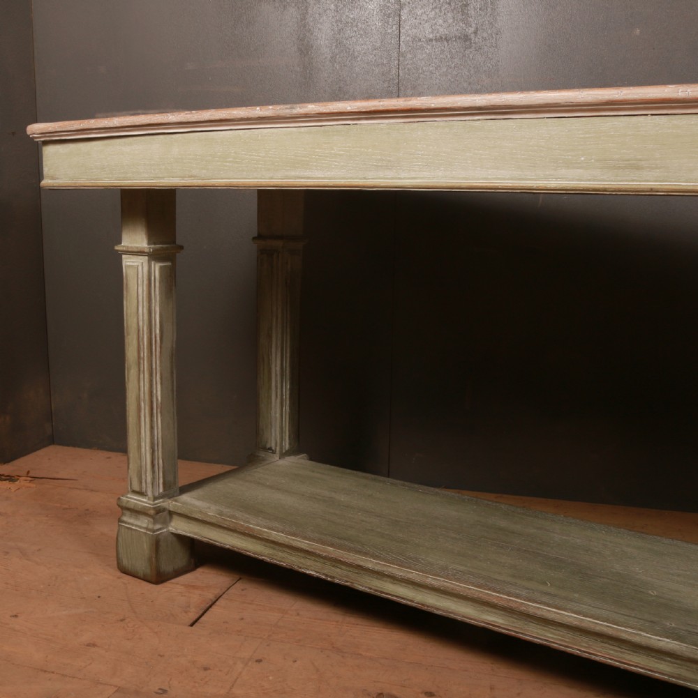 A long French drapers table featuring a muted sage green painted frame with six fluted legs, a lower full-length shelf, and a light natural timber plank top. View III