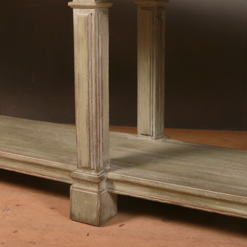 A long French drapers table featuring a muted sage green painted frame with six fluted legs, a lower full-length shelf, and a light natural timber plank top. View V