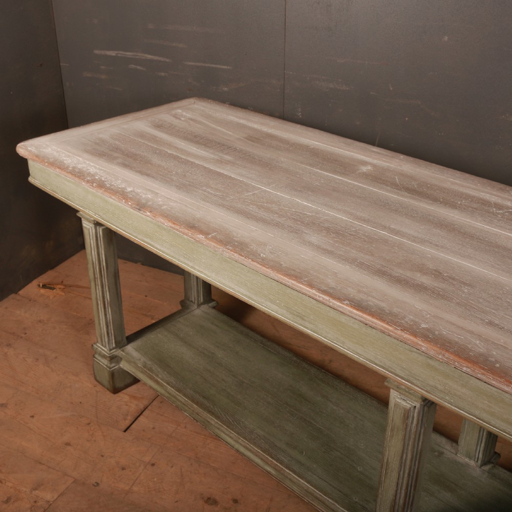 A long French drapers table featuring a muted sage green painted frame with six fluted legs, a lower full-length shelf, and a light natural timber plank top. View VI